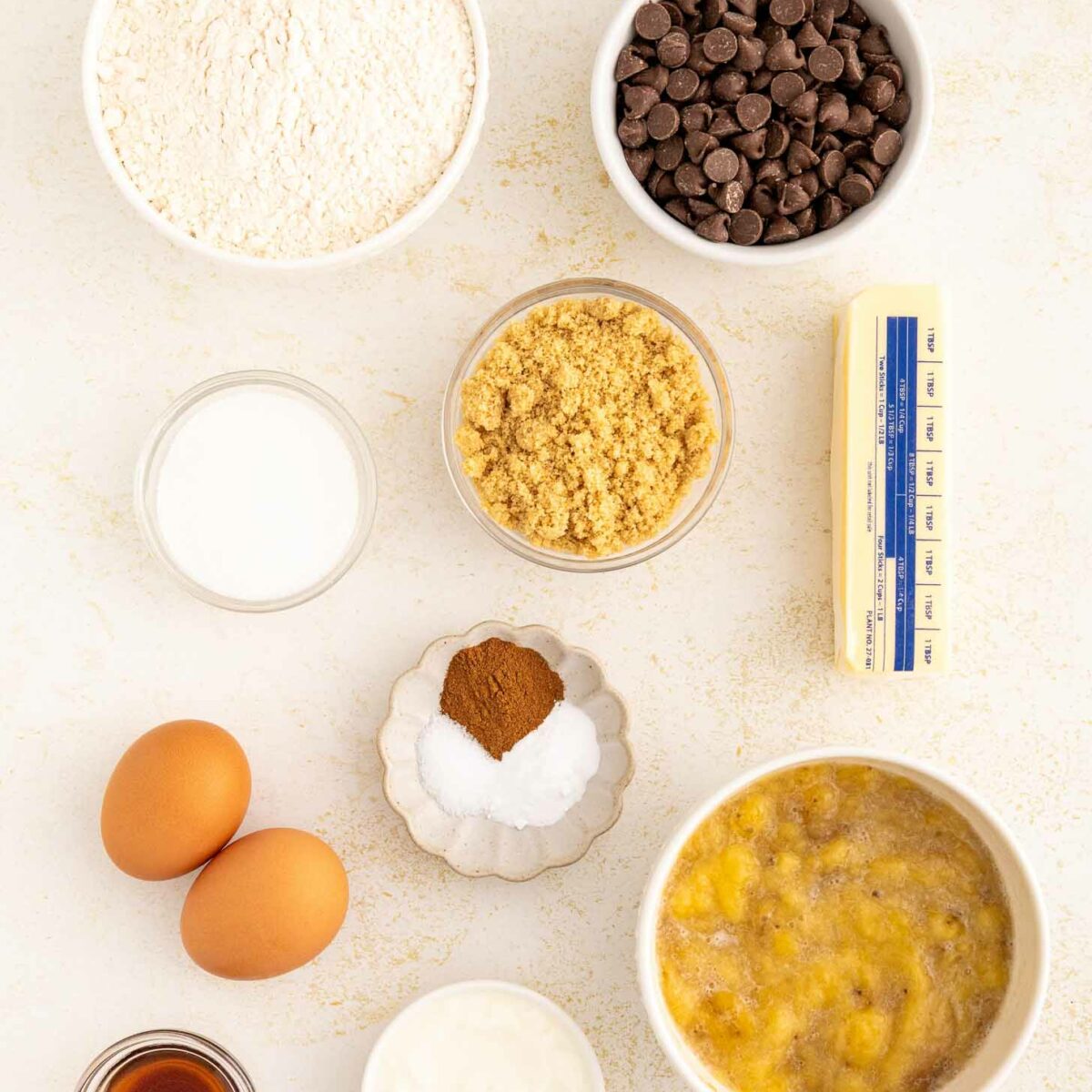 Ingredients for baking are arranged on a white surface, including flour, chocolate chips, brown sugar, butter, granulated sugar, spices, eggs, vanilla extract, yogurt, and a bowl of mashed bananas.