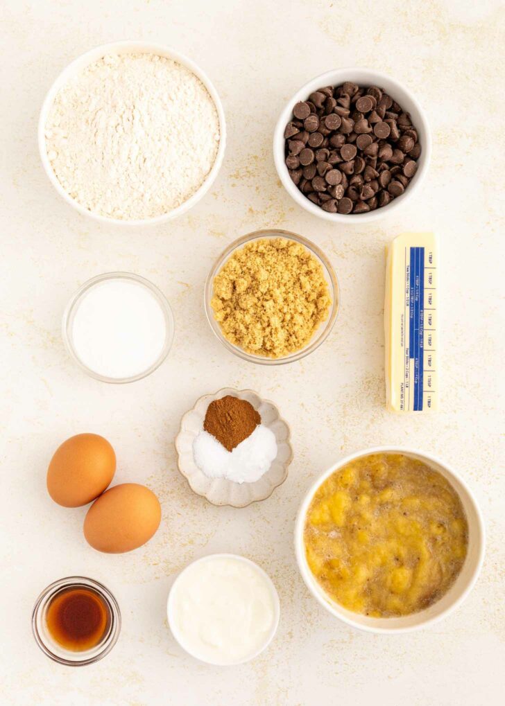 Ingredients for baking are arranged on a white surface, including flour, chocolate chips, brown sugar, butter, granulated sugar, spices, eggs, vanilla extract, yogurt, and a bowl of mashed bananas.
