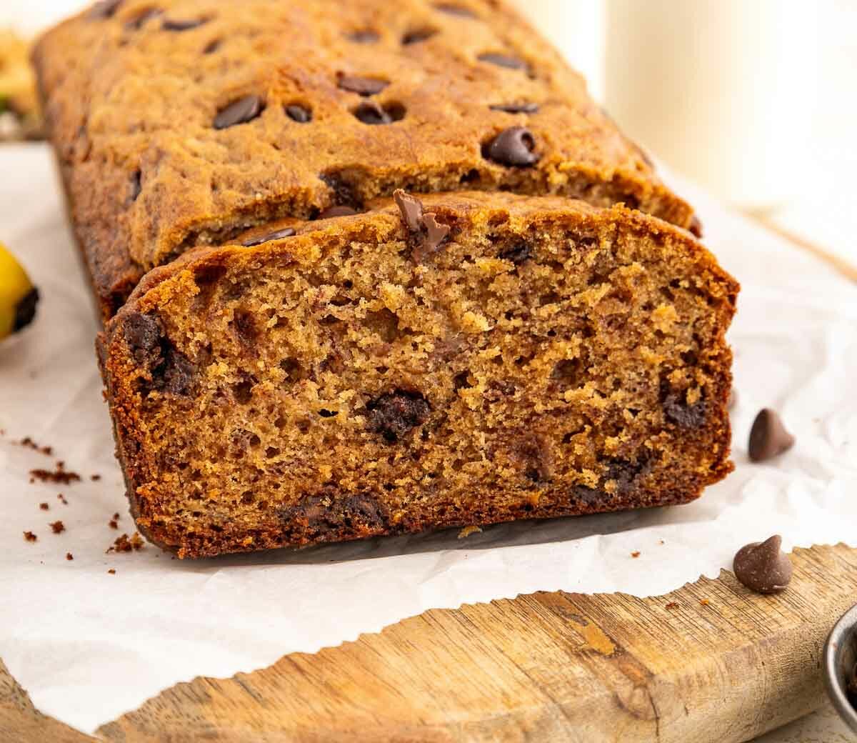 A loaf of chocolate chip banana bread sits on parchment paper atop a wooden board, with one slice cut to show its moist texture and chocolate chips inside. There are chocolate chips and a banana nearby.