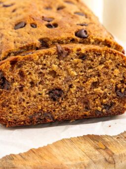A loaf of chocolate chip banana bread sits on parchment paper atop a wooden board, with one slice cut to show its moist texture and chocolate chips inside. There are chocolate chips and a banana nearby.