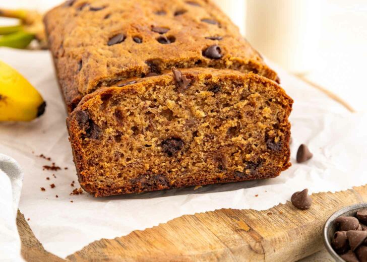 A loaf of chocolate chip banana bread sits on parchment paper atop a wooden board, with one slice cut to show its moist texture and chocolate chips inside. There are chocolate chips and a banana nearby.