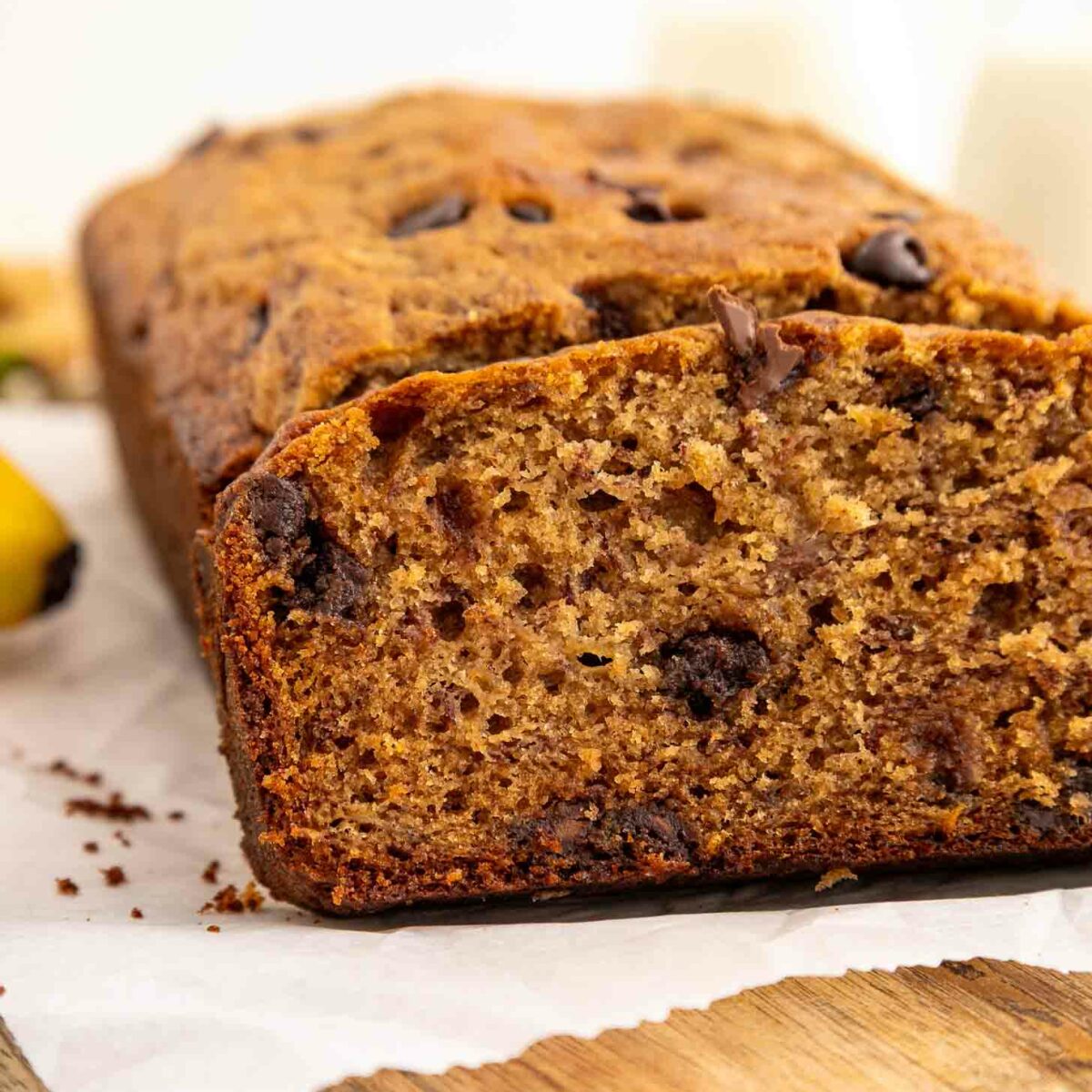 A close-up of a sliced loaf of banana bread with visible chocolate chips, resting on parchment paper atop a wooden surface. The bread looks moist and golden brown.