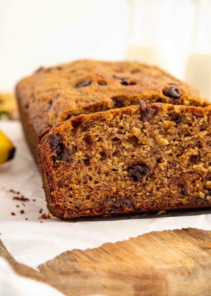 A close-up of a sliced loaf of banana bread with visible chocolate chips, resting on parchment paper atop a wooden surface. The bread looks moist and golden brown.