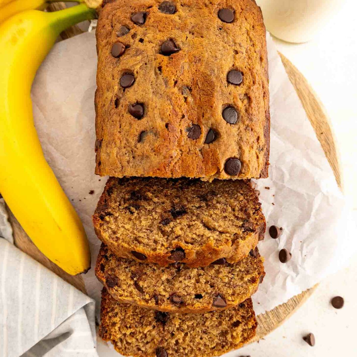 A loaf of chocolate chip banana bread, partially sliced, sits on parchment paper with whole bananas, scattered chocolate chips, and two glasses of milk nearby on a light surface.