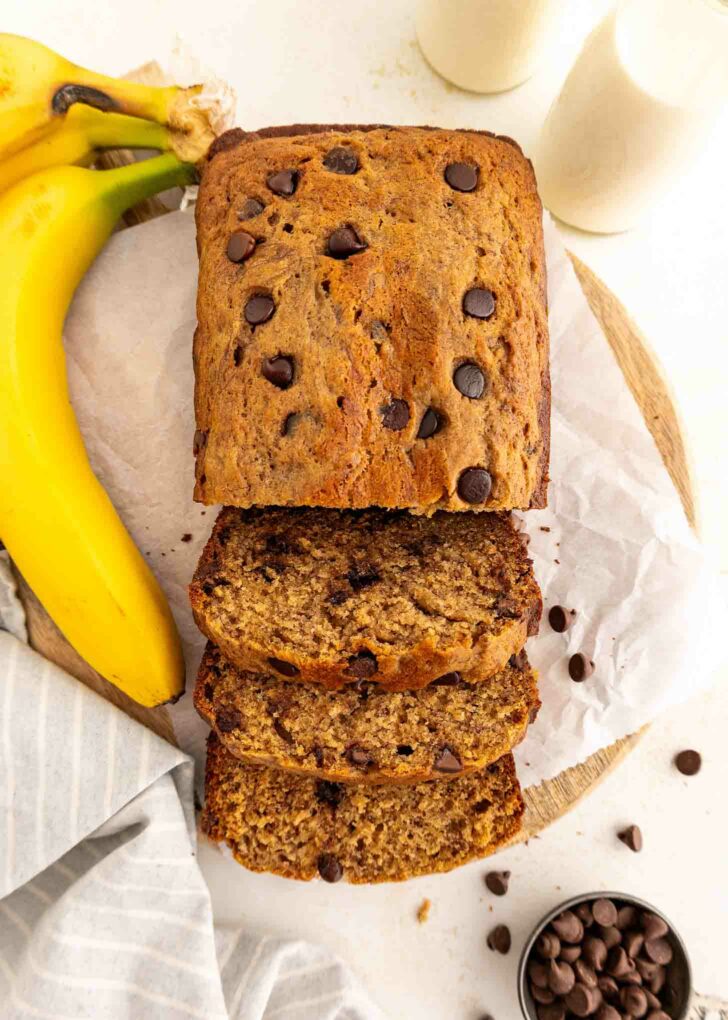 A loaf of chocolate chip banana bread, partially sliced, sits on parchment paper with whole bananas, scattered chocolate chips, and two glasses of milk nearby on a light surface.