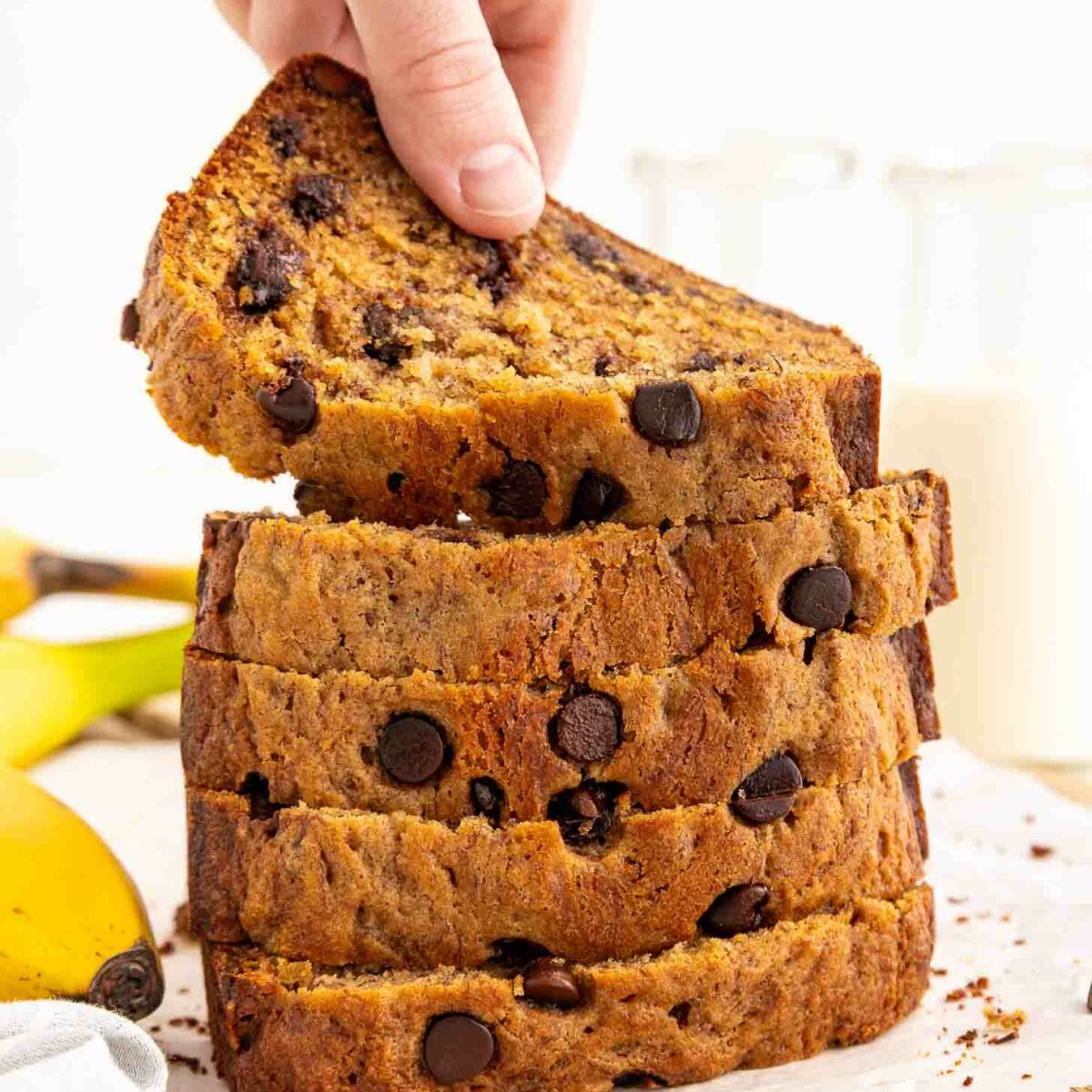 A hand lifts a slice from a stack of thick, chocolate chip-filled banana bread slices on parchment paper. Bananas and glass bottles of milk are visible in the background.