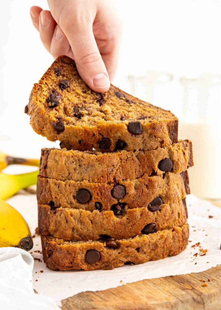 A hand lifts a slice from a stack of thick, chocolate chip-filled banana bread slices on parchment paper. Bananas and glass bottles of milk are visible in the background.