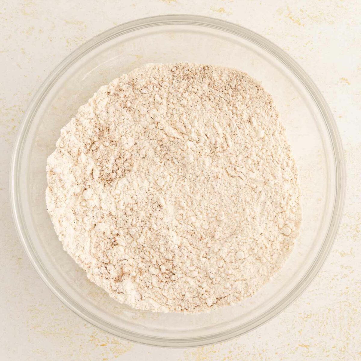 A clear glass bowl filled with a flour mixture sits on a light, textured surface.