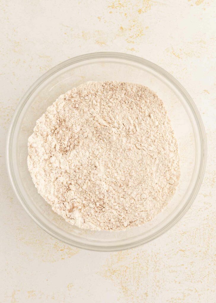 A clear glass bowl filled with a flour mixture sits on a light, textured surface.