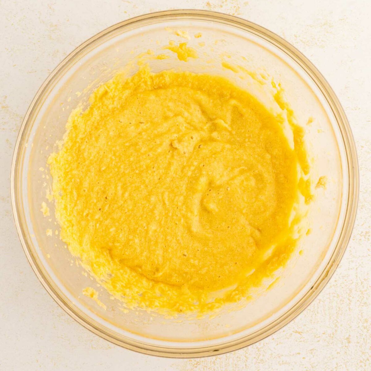 A clear glass bowl filled with a yellow, slightly lumpy batter sits on a light-colored countertop.