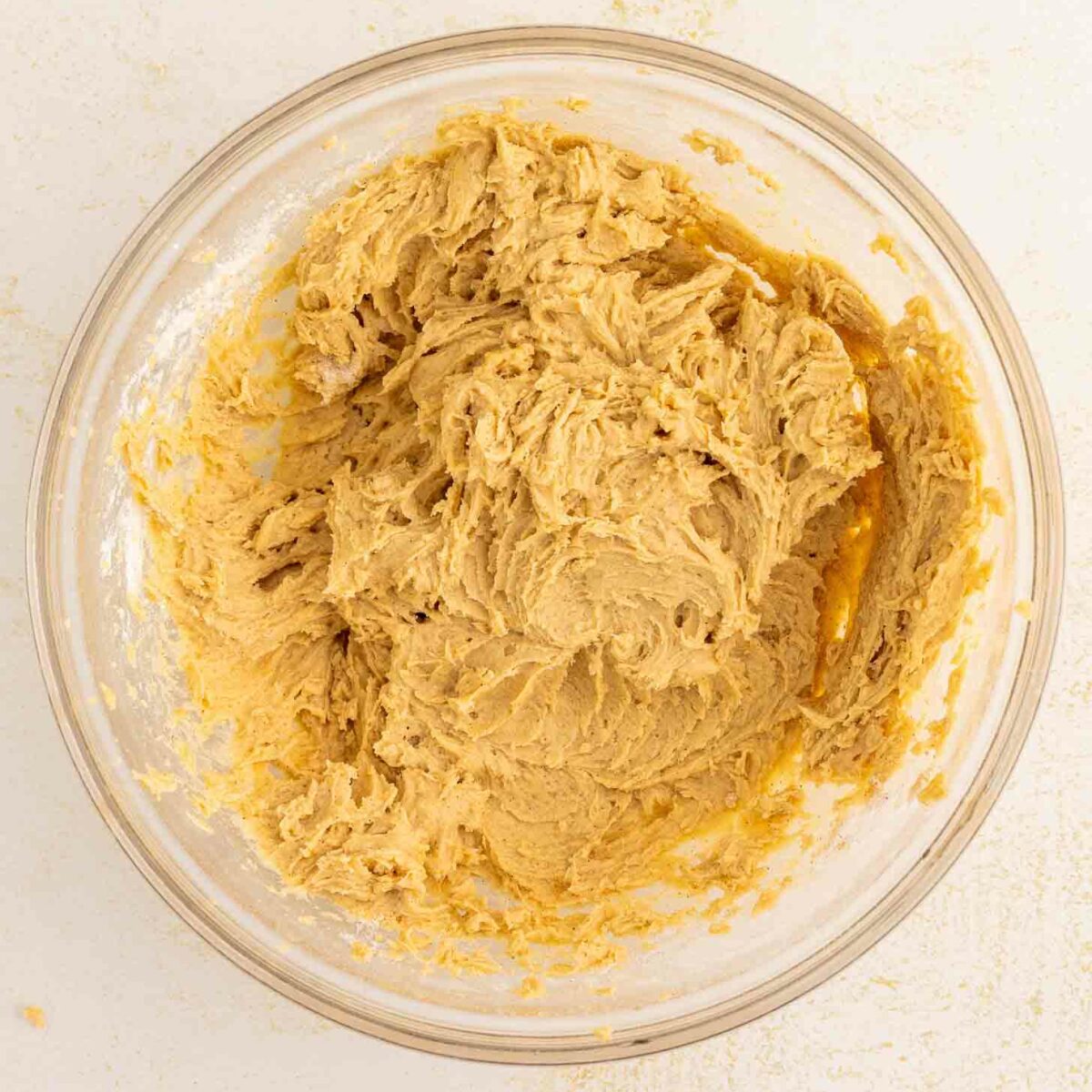 A clear glass bowl filled with light brown  dough mixture sits on a white surface. Some dough is smeared along the sides of the bowl.