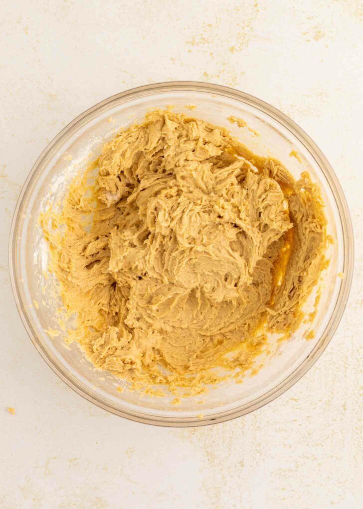 A clear glass bowl filled with light brown  dough mixture sits on a white surface. Some dough is smeared along the sides of the bowl.