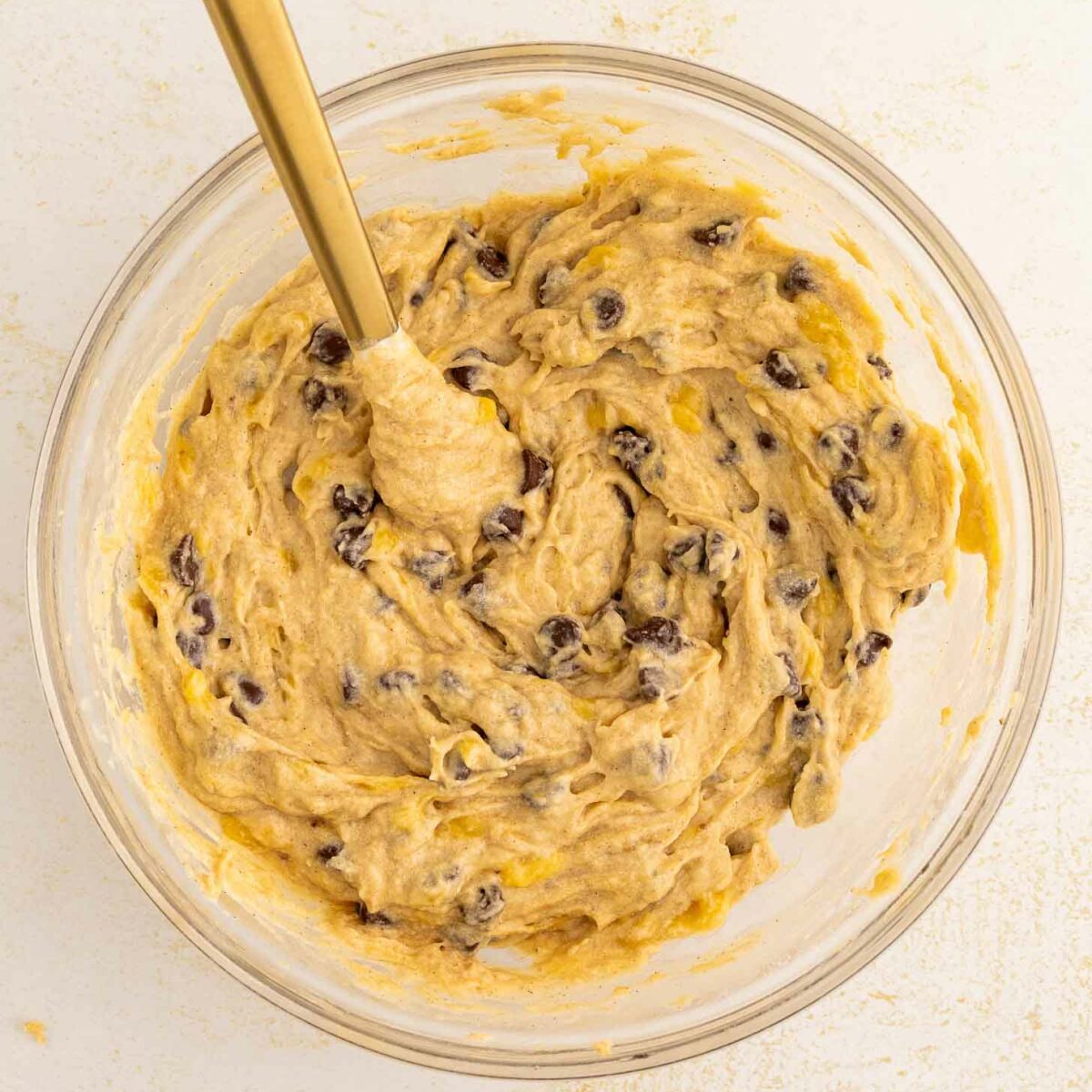 A glass bowl filled with chocolate chip banana bread dough and a gold spoon resting inside, on a light-colored surface.