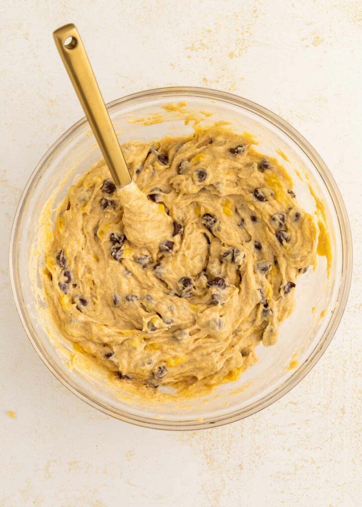 A glass bowl filled with chocolate chip banana bread dough and a gold spoon resting inside, on a light-colored surface.