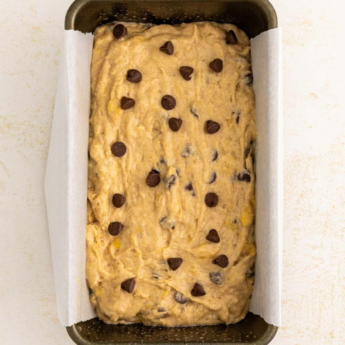 A loaf pan lined with parchment paper, filled with unbaked banana bread batter topped with chocolate chips, sits on a light-colored surface.