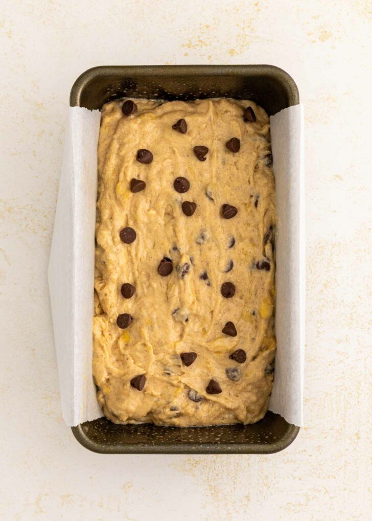 A loaf pan lined with parchment paper, filled with unbaked banana bread batter topped with chocolate chips, sits on a light-colored surface.