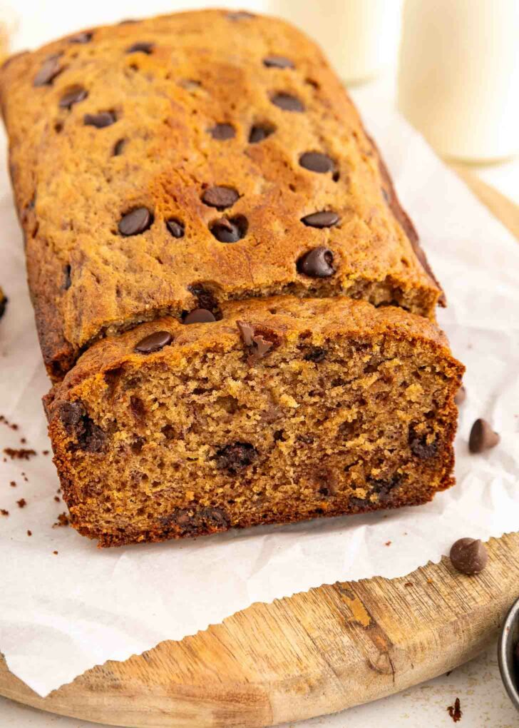 A loaf of banana bread with chocolate chips sits on parchment paper atop a wooden board. One slice has been cut, revealing a moist, dense texture with chocolate chips throughout.