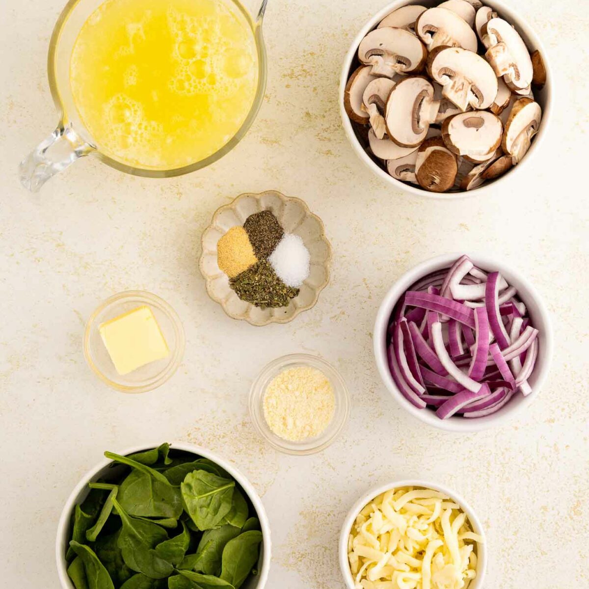 Top-down view of ingredients for a dish: a pitcher of beaten egg whites, bowls of sliced mushrooms, red onion strips, fresh spinach, shredded cheese, a pat of butter, and small dishes of spices and grated cheese.