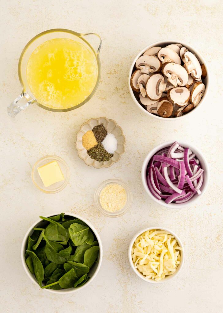 Top-down view of ingredients for a dish: a pitcher of beaten egg whites, bowls of sliced mushrooms, red onion strips, fresh spinach, shredded cheese, a pat of butter, and small dishes of spices and grated cheese.