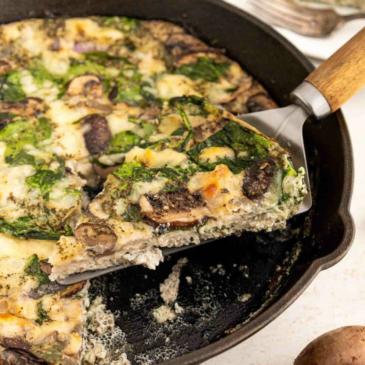 A slice of vegetable frittata with spinach and mushrooms is being lifted from a cast-iron skillet. The frittata is creamy and dotted with greens and mushrooms, with another slice on a plate in the background.