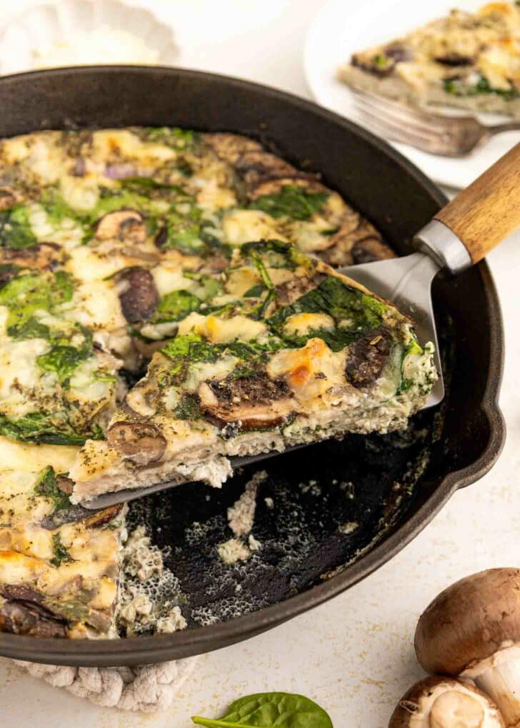 A slice of vegetable frittata with spinach and mushrooms is being lifted from a cast-iron skillet. The frittata is creamy and dotted with greens and mushrooms, with another slice on a plate in the background.
