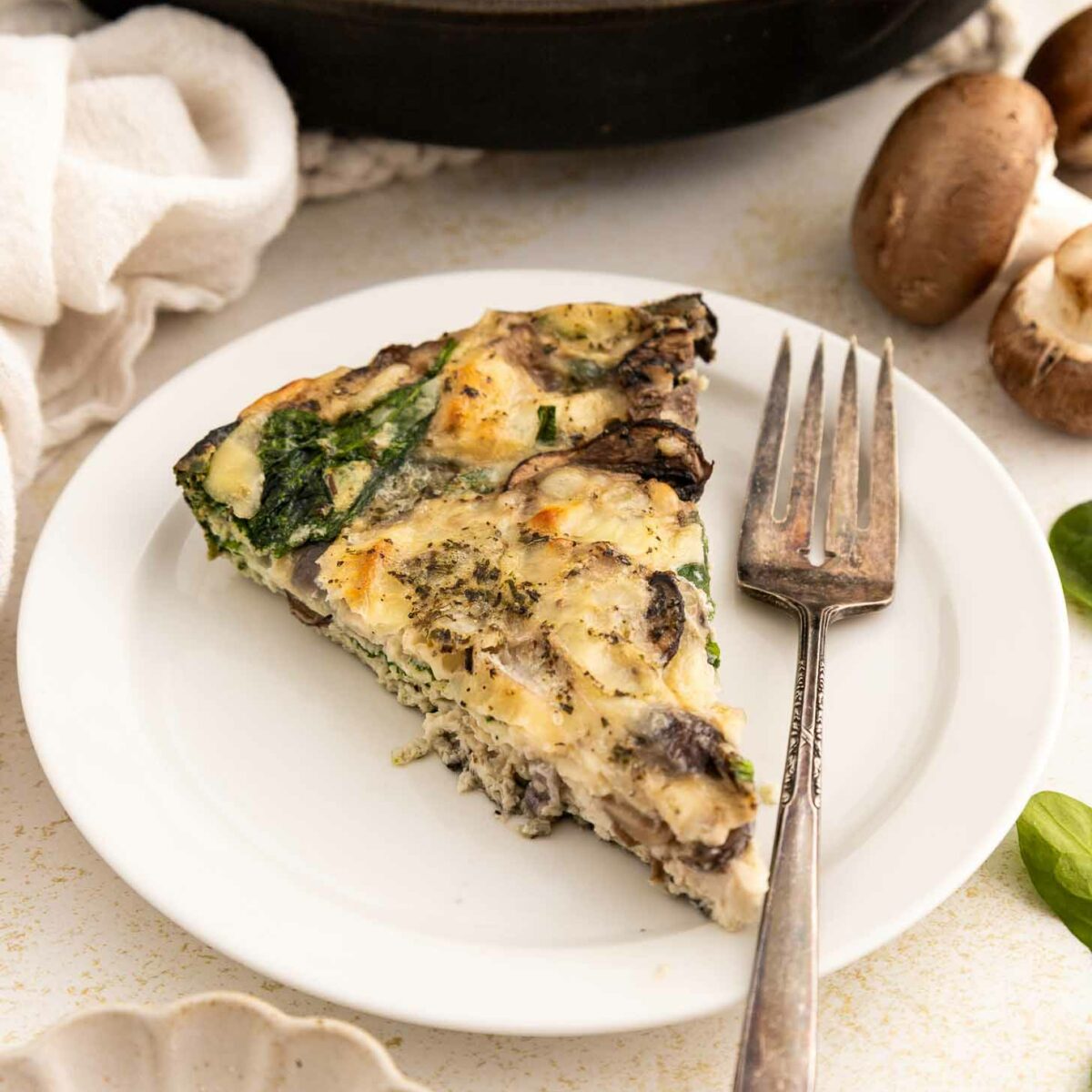 A slice of spinach and mushroom egg white frittata on a white plate with a fork, next to fresh mushrooms, greens, and a dish of grated cheese. The remaining frittata is visible in a skillet in the background.