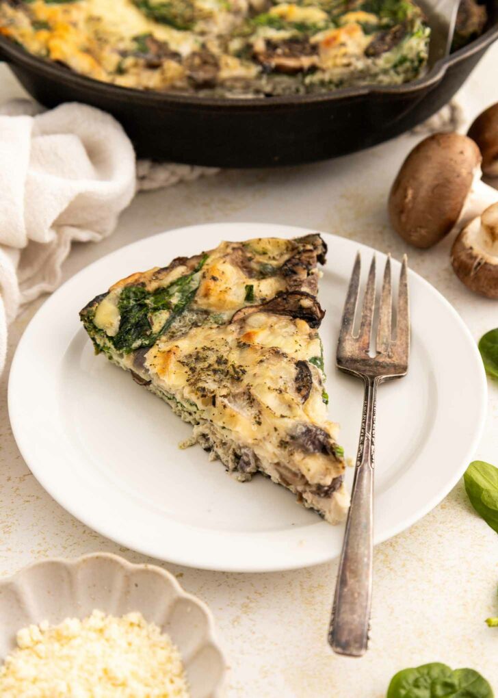 A slice of spinach and mushroom egg white frittata on a white plate with a fork, next to fresh mushrooms, greens, and a dish of grated cheese. The remaining frittata is visible in a skillet in the background.