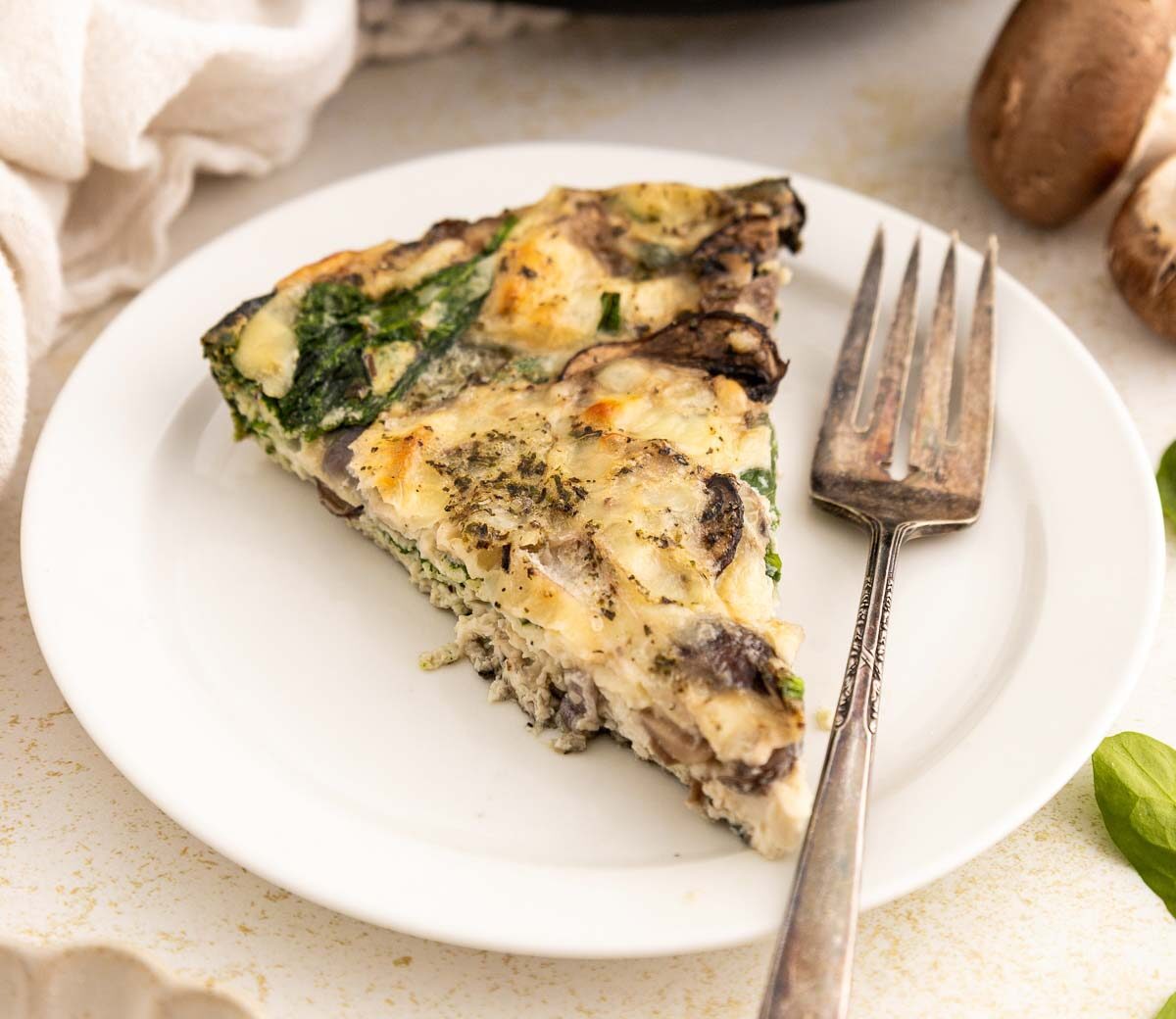 A slice of egg white frittata with mushrooms, spinach, and herbs is served on a white plate with a fork. Fresh spinach leaves and mushrooms are visible nearby on the light-colored table.