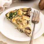 A slice of egg white frittata with mushrooms, spinach, and herbs is served on a white plate with a fork. Fresh spinach leaves and mushrooms are visible nearby on the light-colored table.