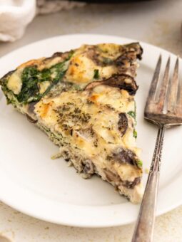A slice of egg white frittata with mushrooms, spinach, and herbs is served on a white plate with a fork. Fresh spinach leaves and mushrooms are visible nearby on the light-colored table.