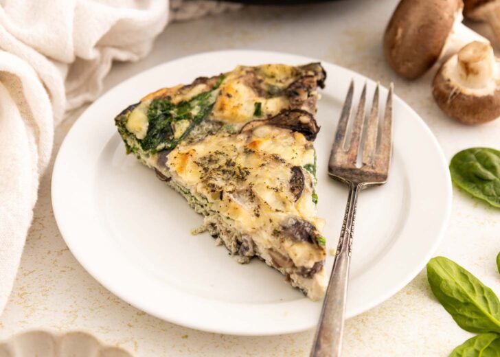 A slice of egg white frittata with mushrooms, spinach, and herbs is served on a white plate with a fork. Fresh spinach leaves and mushrooms are visible nearby on the light-colored table.