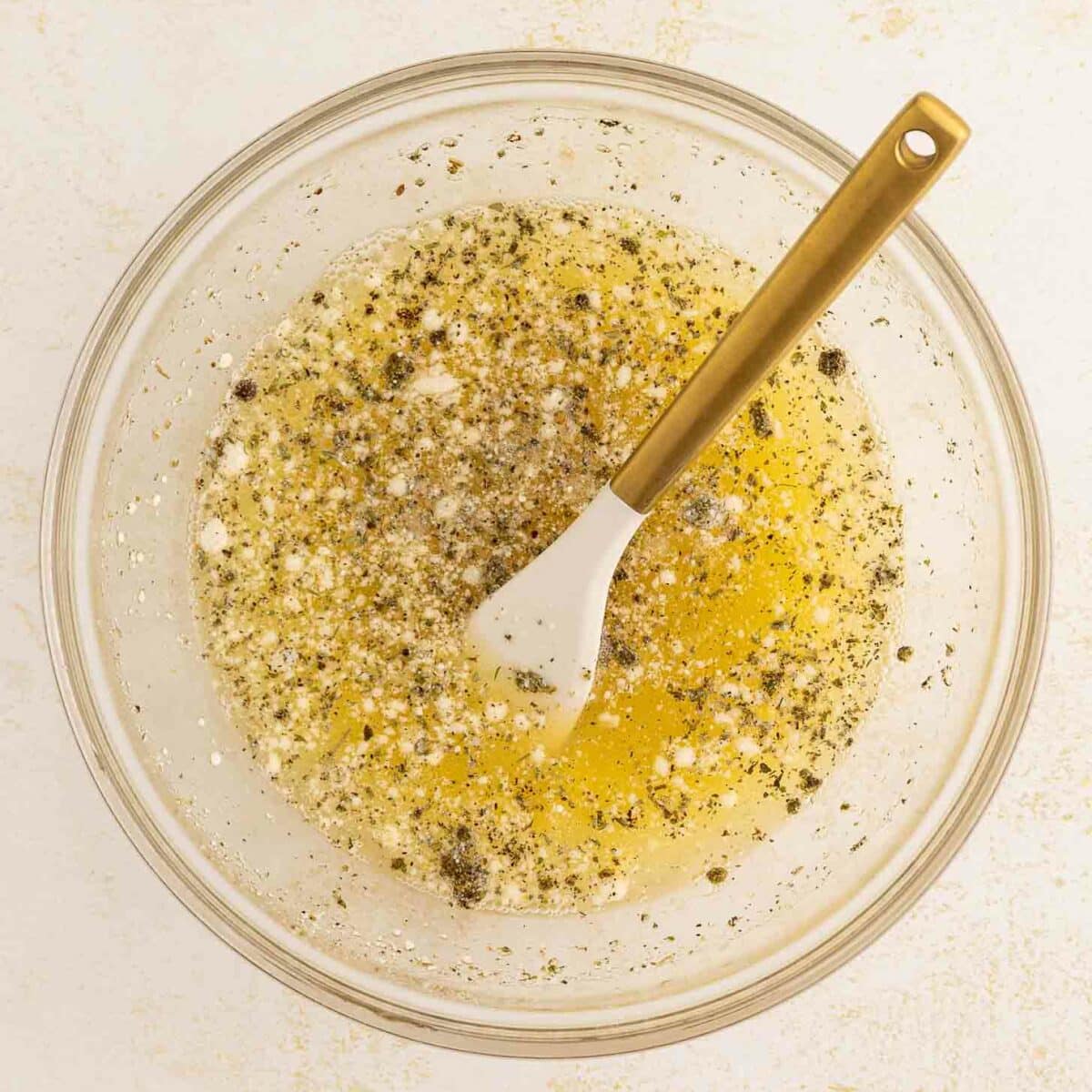 A glass bowl filled with egg whites, seasonings, and cracked black pepper, with a white spatula featuring a gold handle resting inside. The bowl is placed on a light-colored surface.