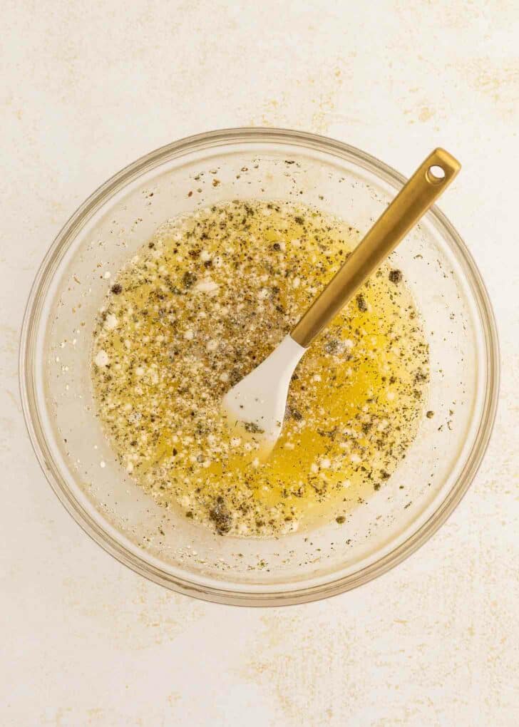 A glass bowl filled with egg whites, seasonings, and cracked black pepper, with a white spatula featuring a gold handle resting inside. The bowl is placed on a light-colored surface.