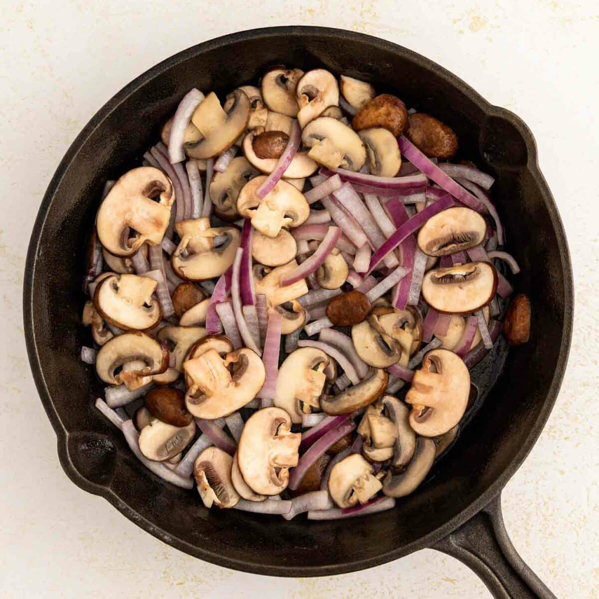 A cast-iron skillet filled with sliced mushrooms and thinly sliced red onions, ready to be cooked, on a light-colored surface.