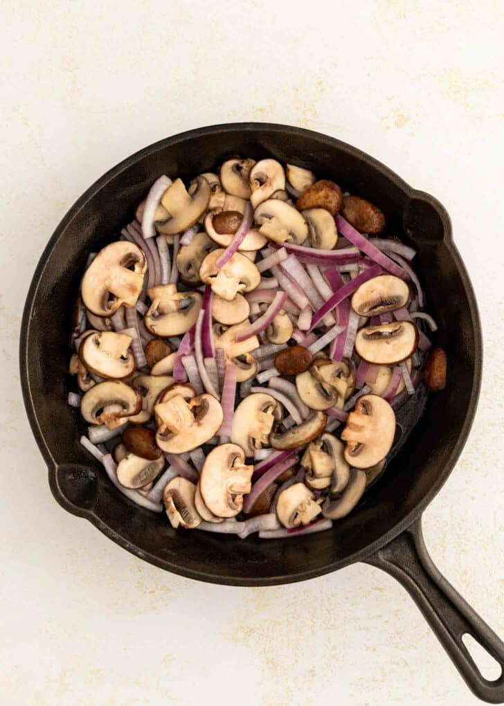 A cast-iron skillet filled with sliced mushrooms and thinly sliced red onions, ready to be cooked, on a light-colored surface.