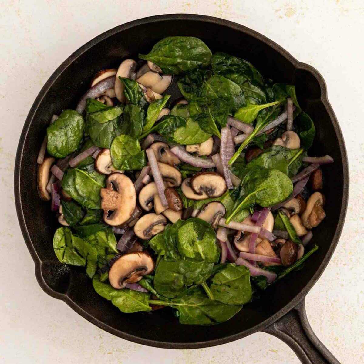 A black skillet filled with sautéed spinach, sliced mushrooms, and red onions on a light-colored surface.