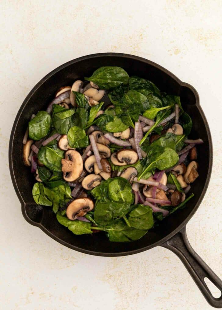 A black skillet filled with sautéed spinach, sliced mushrooms, and red onions on a light-colored surface.