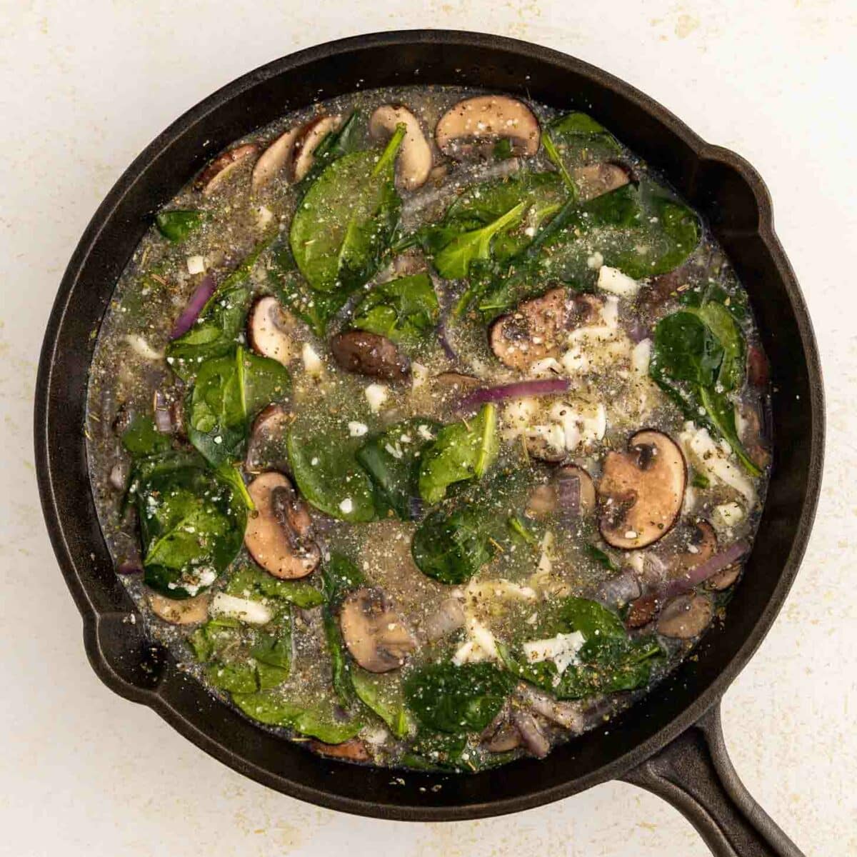 A black skillet filled with a mixture of spinach leaves, sliced mushrooms, onions, and egg whites on a light-colored surface.