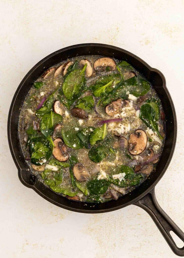 A black skillet filled with a mixture of spinach leaves, sliced mushrooms, onions, and egg whites on a light-colored surface.