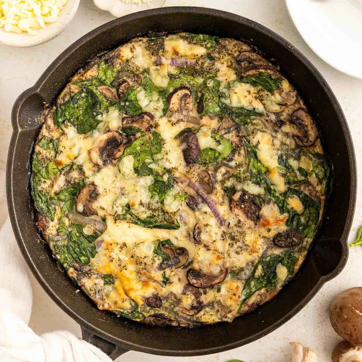 A spinach and mushroom egg white frittata baked in a black skillet, garnished with cheese and herbs. Surrounding the skillet are a fork, small bowls of cheese, mushrooms, and fresh spinach leaves on a white surface.