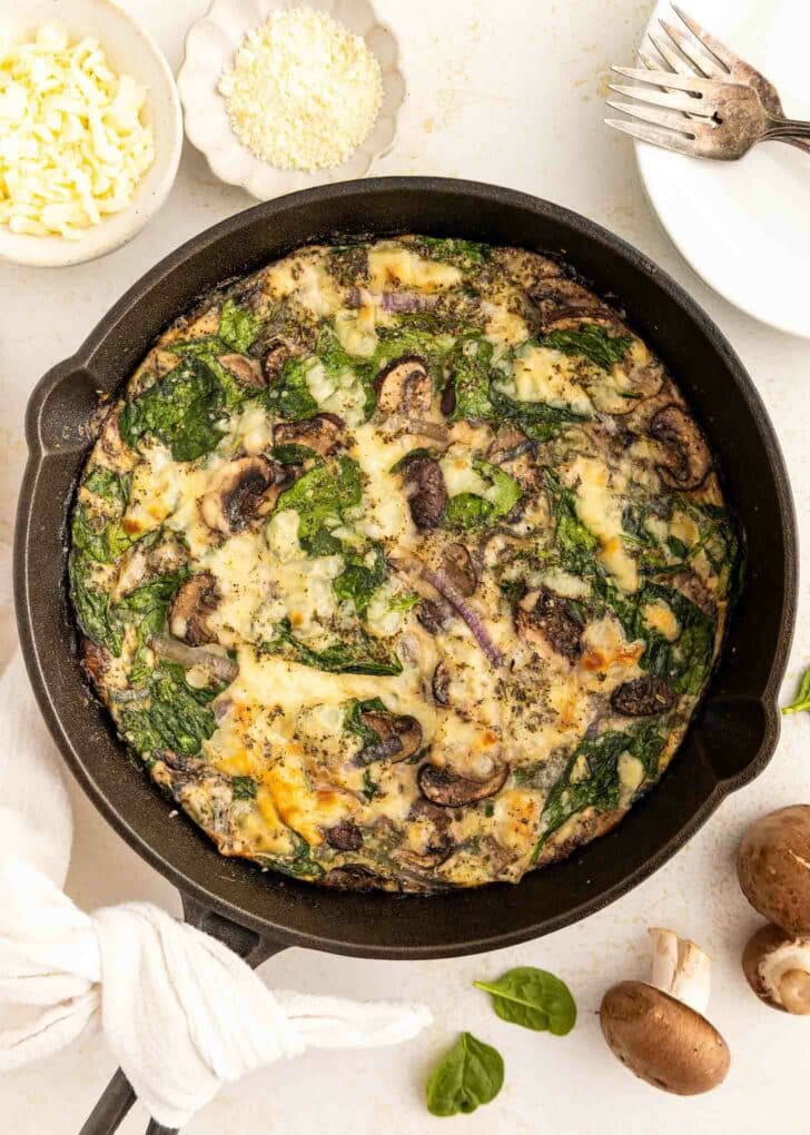 A spinach and mushroom egg white frittata baked in a black skillet, garnished with cheese and herbs. Surrounding the skillet are a fork, small bowls of cheese, mushrooms, and fresh spinach leaves on a white surface.