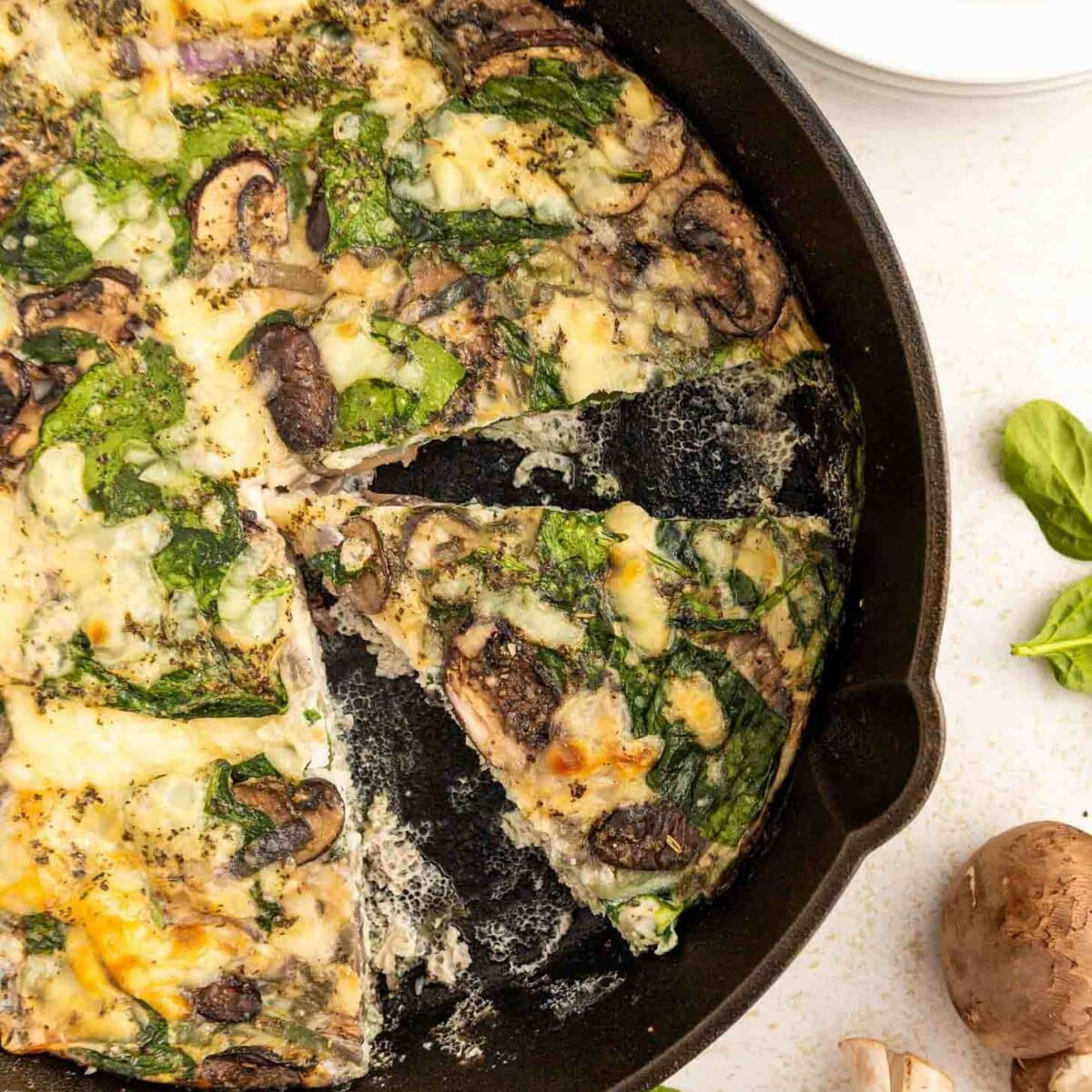 A skillet filled with a sliced mushroom and spinach egg white frittata. One wedge is being removed, and fresh mushrooms and spinach leaves are scattered nearby on a white surface. Plates and a fork are in the background.