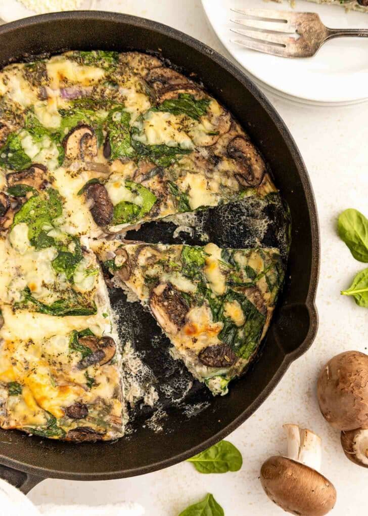 A skillet filled with a sliced mushroom and spinach egg white frittata. One wedge is being removed, and fresh mushrooms and spinach leaves are scattered nearby on a white surface. Plates and a fork are in the background.
