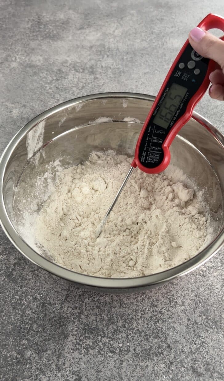 A hand holds a digital thermometer in a mixing bowl of flour, measuring the temperature of the dry ingredients on a gray countertop.