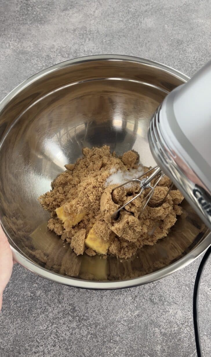 A hand holds a metal mixing bowl containing brown sugar, white sugar, and butter. An electric hand mixer is positioned above the bowl, ready to blend the ingredients.
