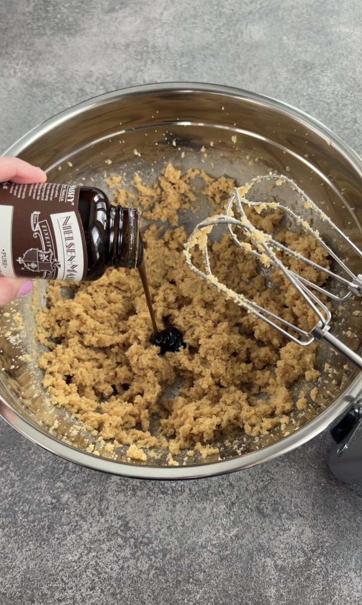 A hand pours vanilla extract from a bottle into a mixing bowl filled with creamed cookie dough. An electric hand mixer with beaters is resting in the bowl. The background is a gray countertop.