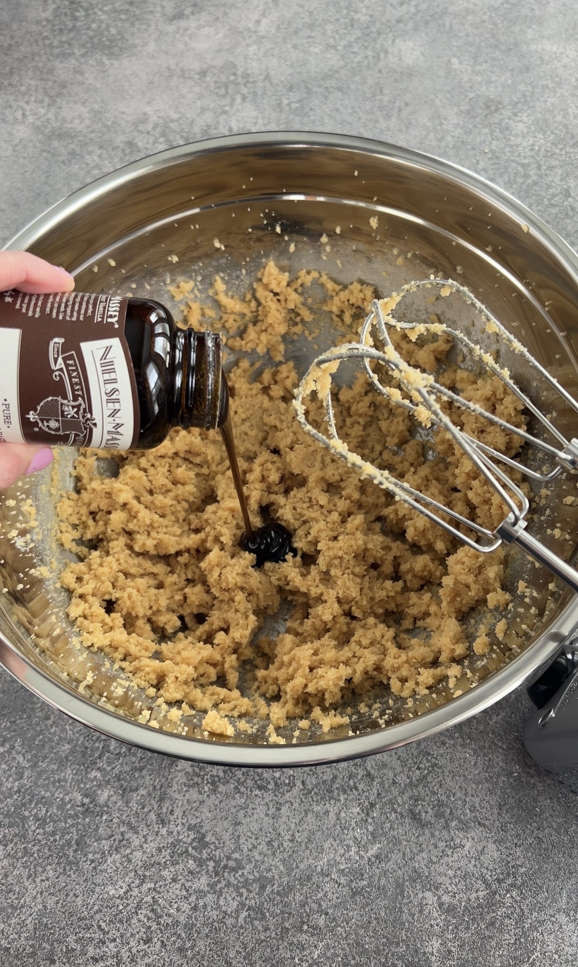A hand pours vanilla extract from a bottle into a mixing bowl filled with creamed cookie dough. An electric hand mixer with beaters is resting in the bowl. The background is a gray countertop.