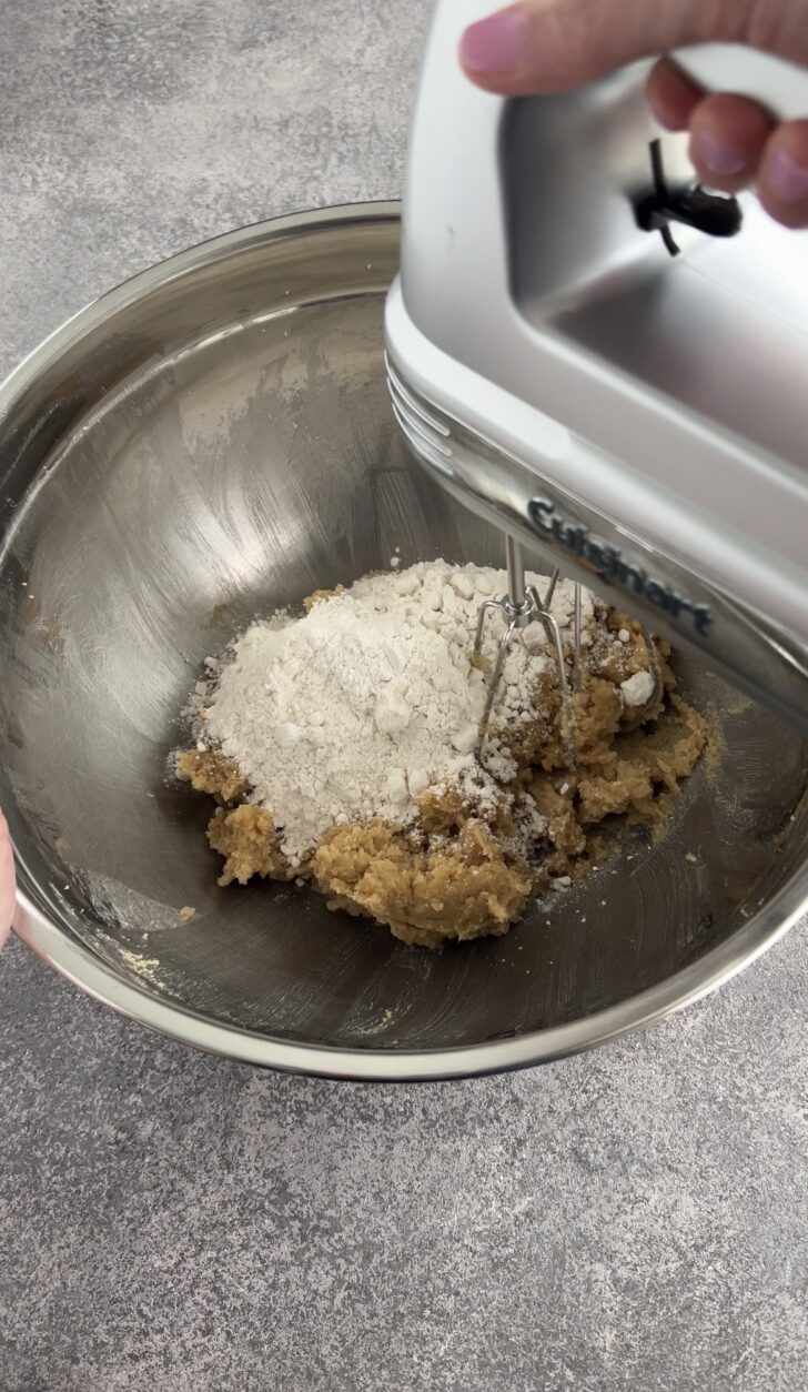 A close-up of hands using an electric hand mixer to blend flour into cookie dough in a large stainless steel mixing bowl on a gray countertop.