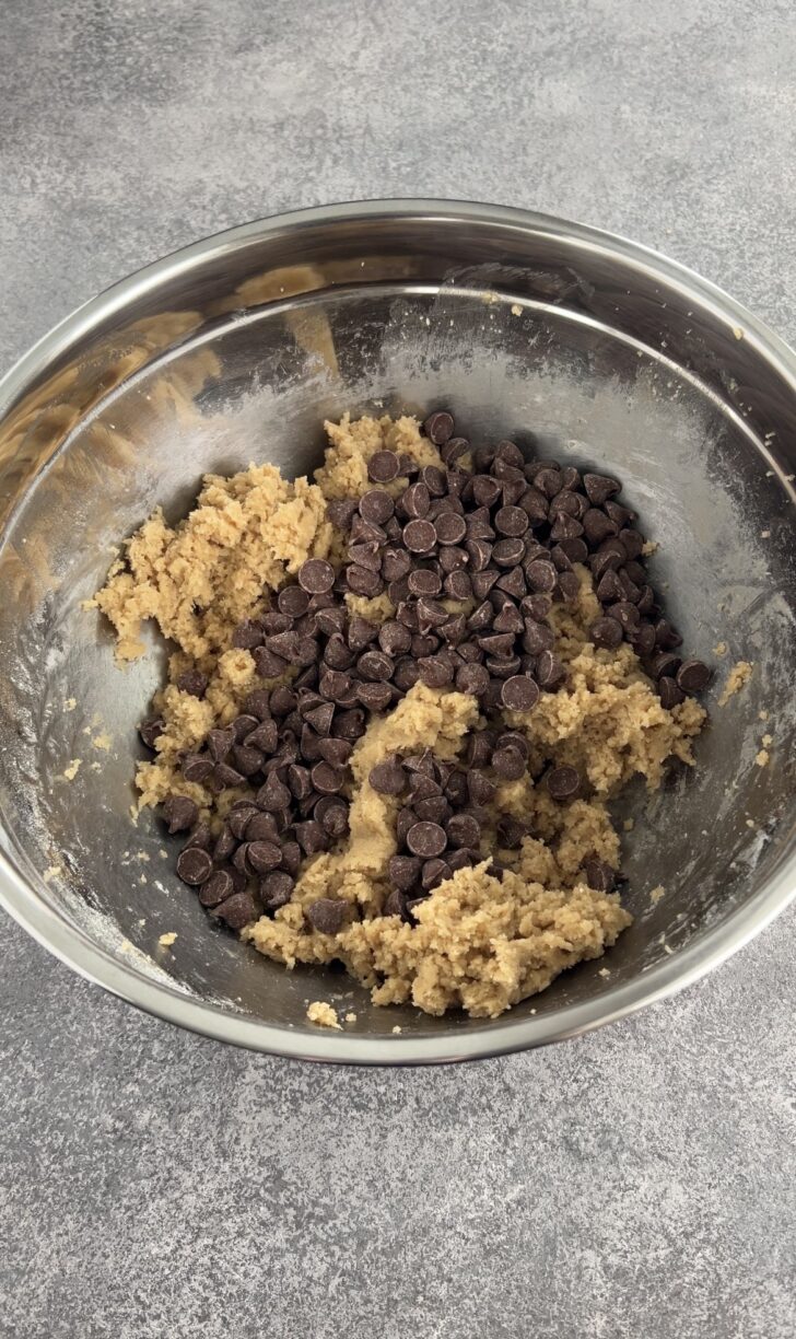 A metal mixing bowl containing cookie dough and a generous amount of chocolate chips on top, set on a gray countertop.