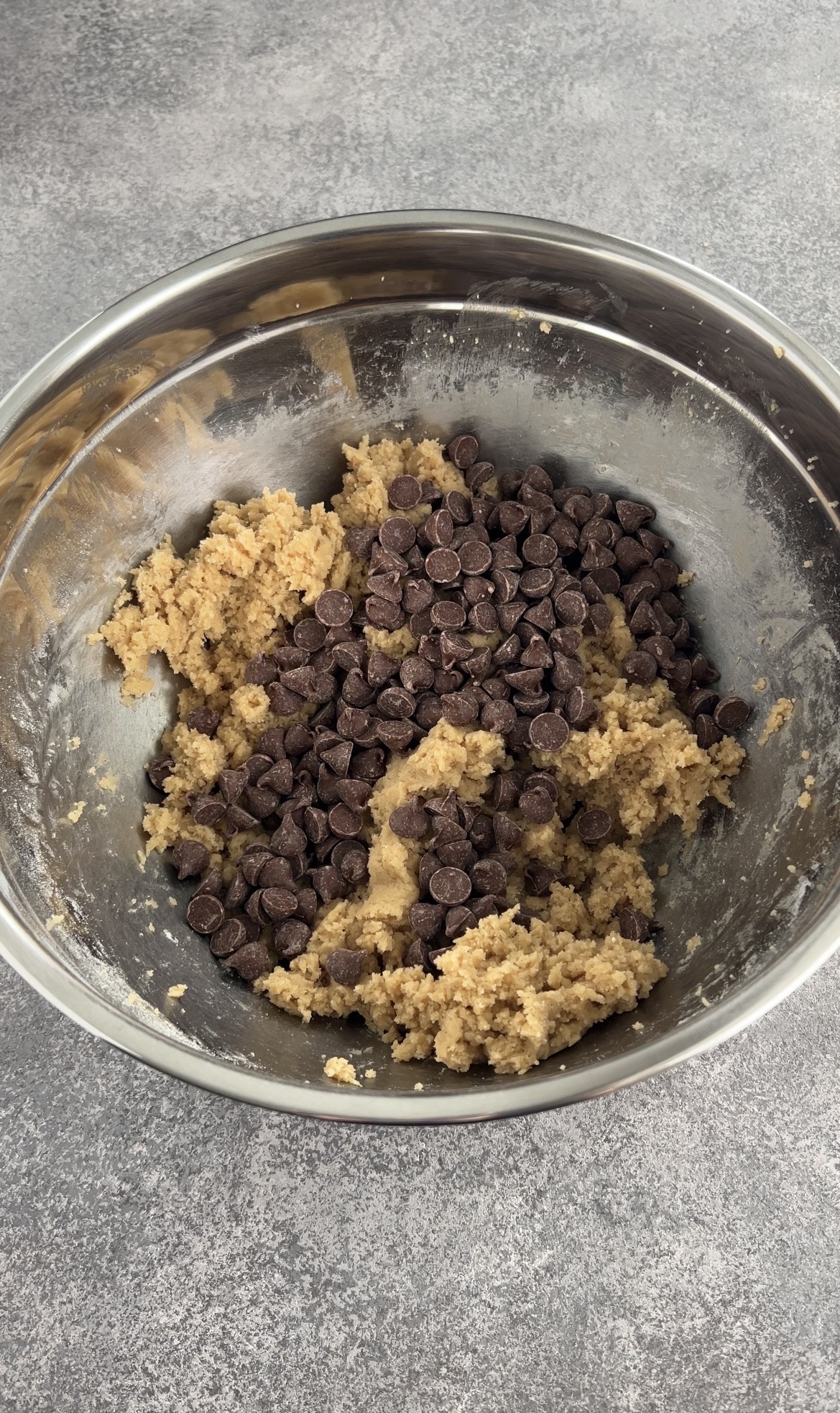 A metal mixing bowl containing cookie dough and a generous amount of chocolate chips on top, set on a gray countertop.